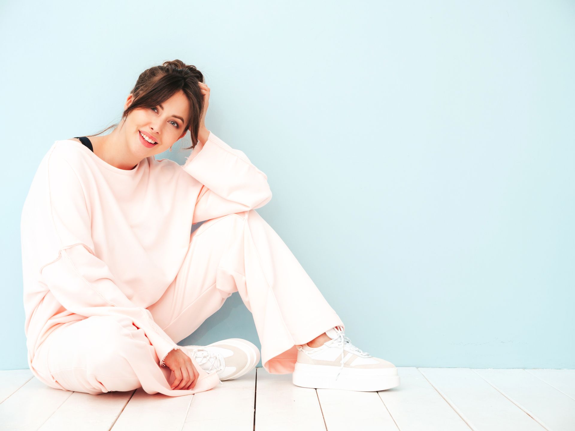 A smiling person in pale pink lounge clothes sits on a white floor against a light blue wall, hand resting on their head.