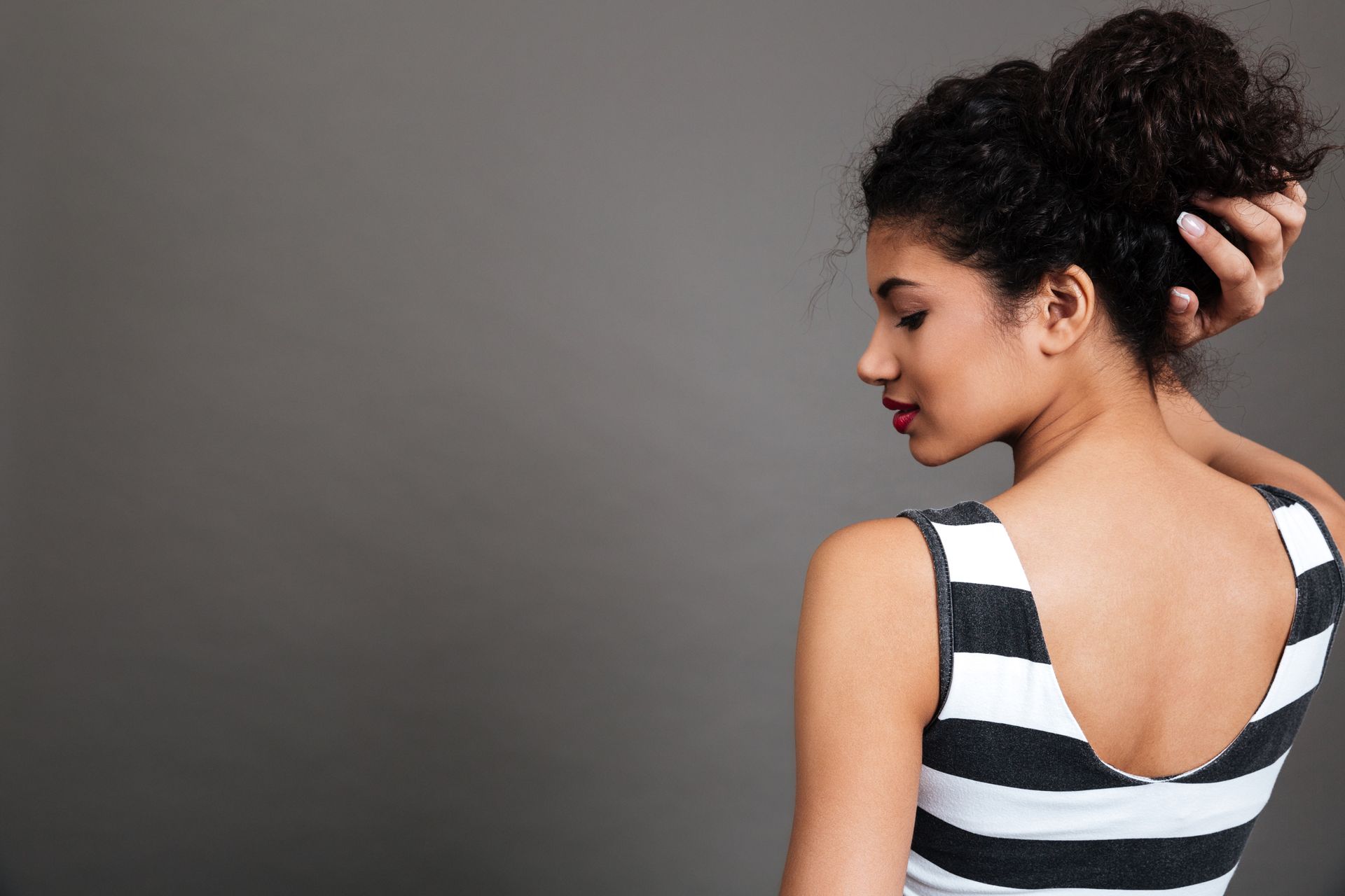 A person with curly hair tied back in a bun, wearing a black-and-white striped tank top, looking down against a grey background.