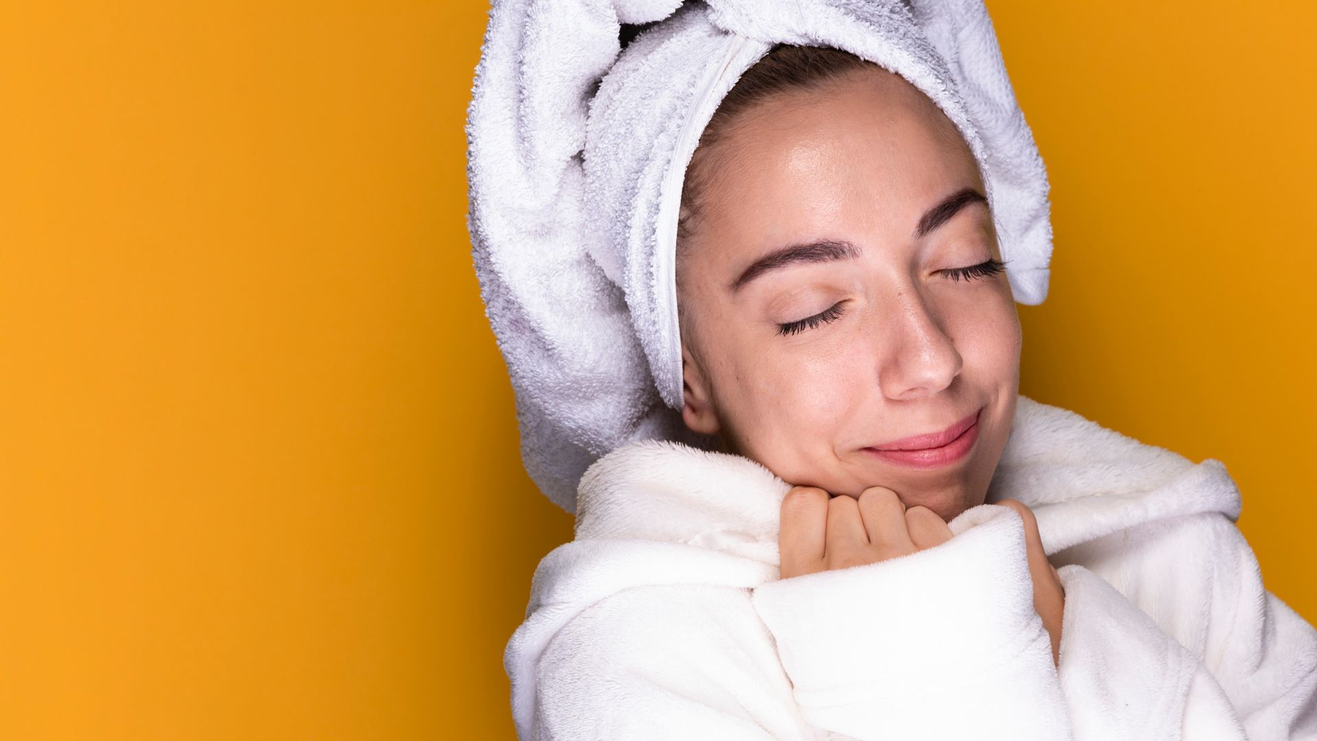 Woman in a white bathrobe and towel, eyes closed, resting against a bright orange background.