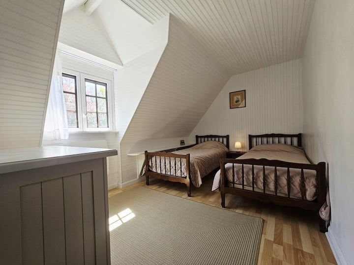 Chambre mansardée lumineuse avec deux lits jumeaux, murs blancs en pente et une fenêtre laissant entrer la lumière du soleil.