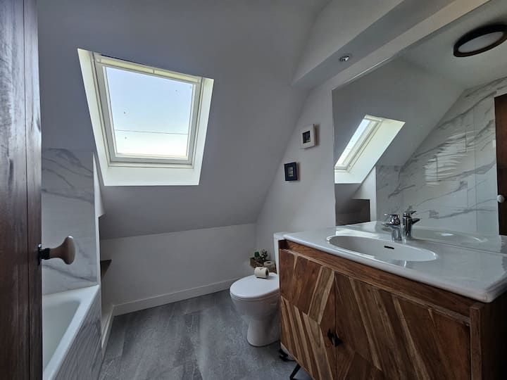 Petite salle de bain mansardée avec puits de lumière, murs blancs, sol gris et meuble-lavabo en bois à côté des toilettes et de la baignoire.