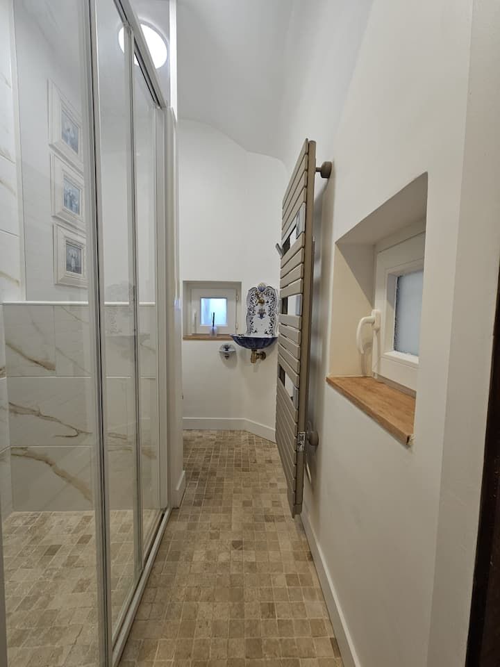 Salle de bains étroite carrelée avec douche en verre, sèche-serviettes et petit rebord de fenêtre avec articles de toilette.