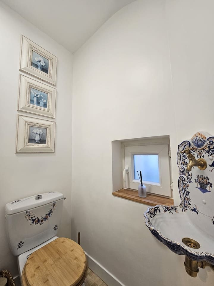 Salle de bains compacte avec murs blancs, toilettes, petit siège en bois, lavabo décoratif et minuscule étagère de fenêtre.