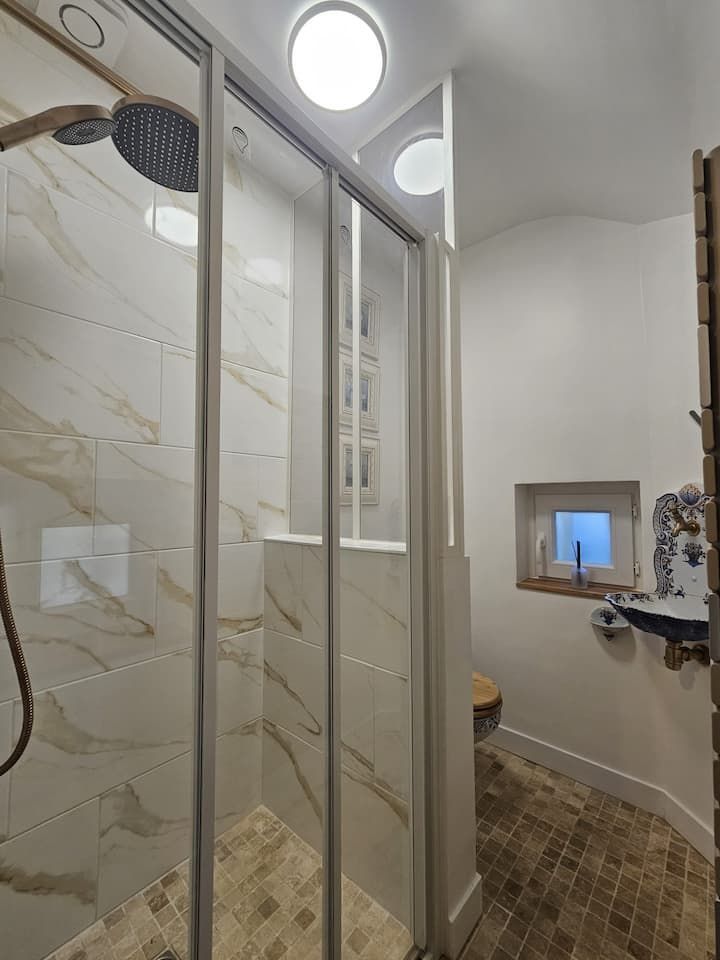 Salle de bains moderne avec paroi de douche en verre, murs imitation marbre et petite fenêtre au-dessus des toilettes.
