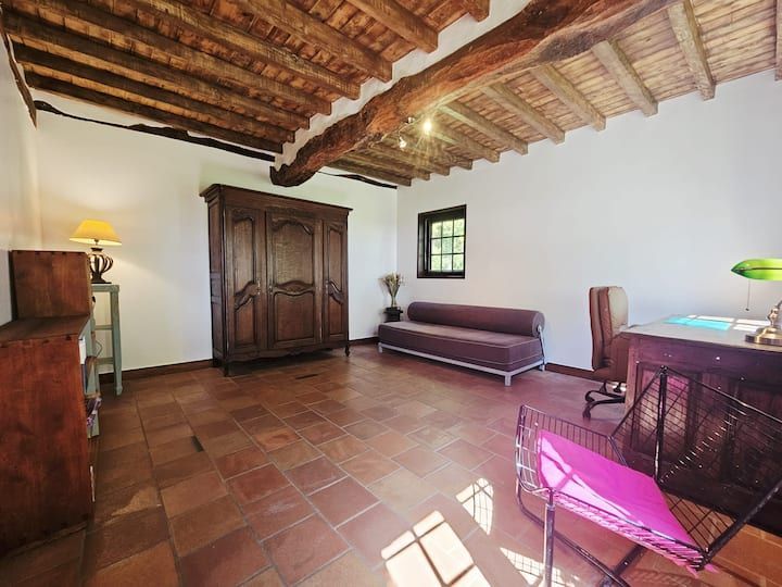 Chambre rustique avec sol en terre cuite, plafond à poutres apparentes, armoire, canapé, bureau et fauteuil rose baigné de lumière naturelle.