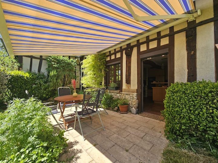 Patio avec auvent rayé, table et chaises en métal, plantes en pot et porte ouverte sur une maison