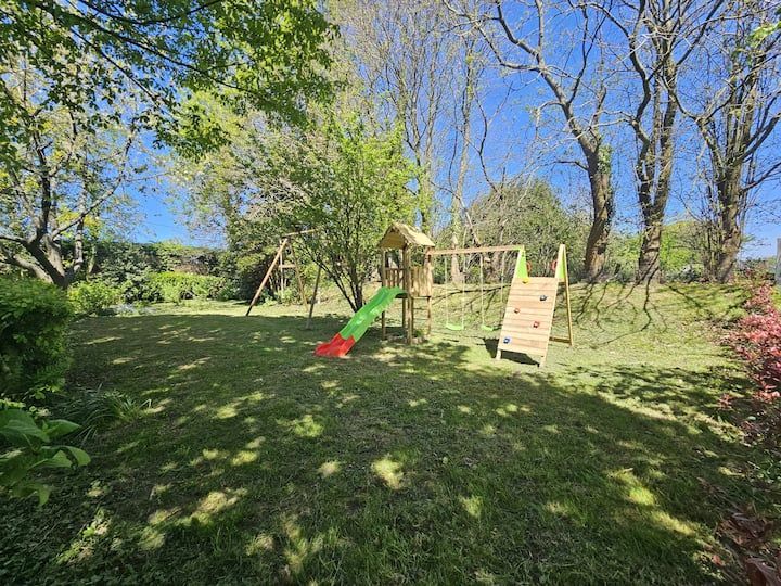 Aire de jeux en bois avec toboggan et mur d'escalade dans le jardin, sur une colline herbeuse sous les arbres et le ciel bleu.