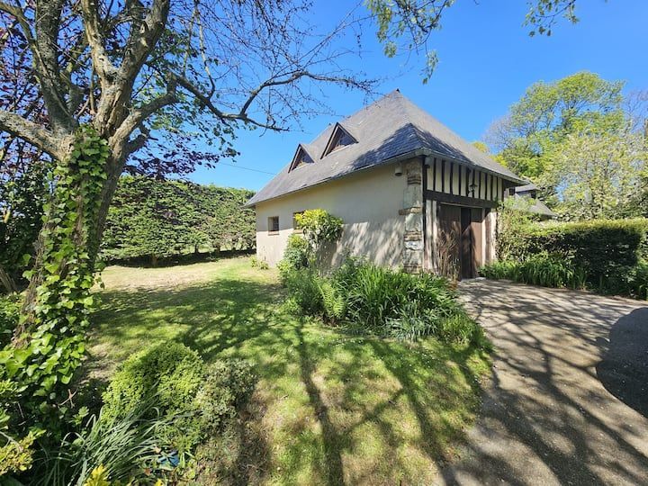 Maison en pierre au toit pentu, située dans un jardin ensoleillé, entourée d'arbres et de haies.