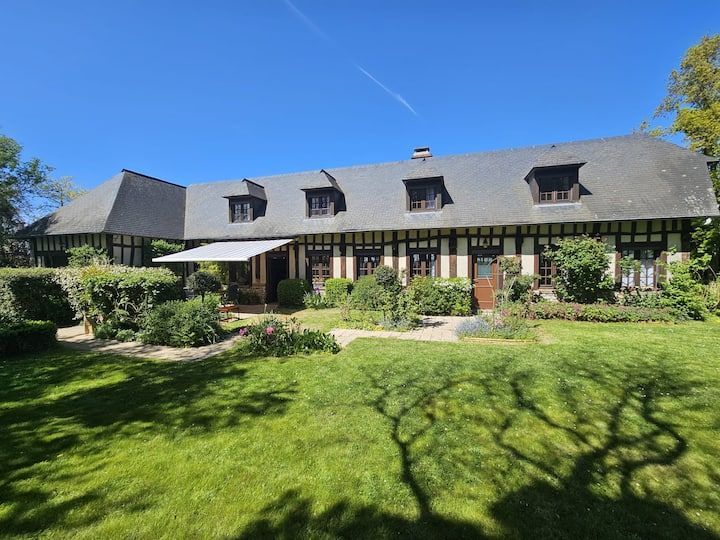 Grande maison en pierre au toit gris, avec porche et pelouse verdoyante sous un ciel d'un bleu éclatant.
