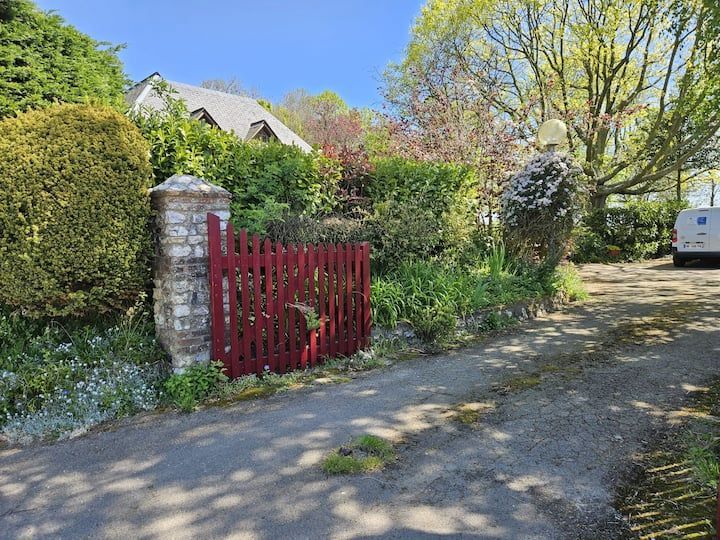 Portail en bois rouge à côté d'un pilier en pierre sur une allée bordée d'arbres, avec une maison et une camionnette garée en arrière-plan.