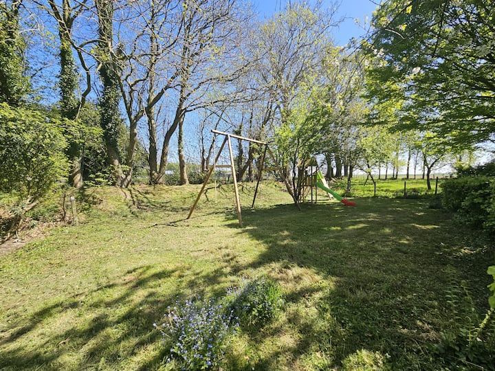 Cour arrière ensoleillée avec pelouse, arbres et balançoire en bois dans un jardin clôturé.