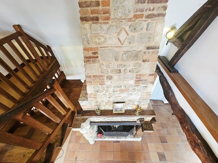 Intérieur rustique avec cheminée en briques, sol carrelé et rampes d'escalier en bois entourant une cage d'escalier ouverte.