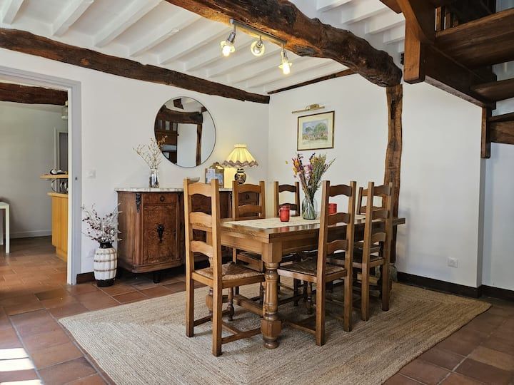 Salle à manger rustique avec table en bois, chaises, buffet, tapis et poutres apparentes au plafond.