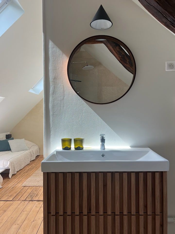 Salle de bain mansardée lumineuse avec meuble-lavabo, miroir rond, applique murale et fenêtres inclinées à côté du lit.