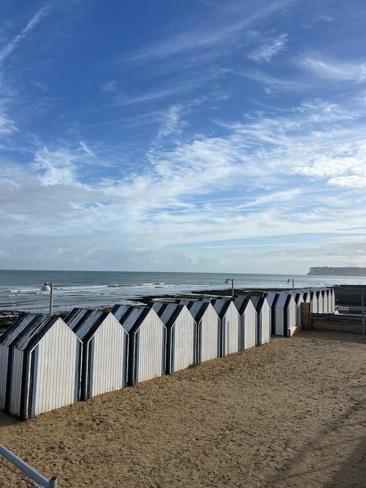 Cabanes de plage sur un rivage sablonneux, avec la mer et un ciel partiellement nuageux en arrière-plan.