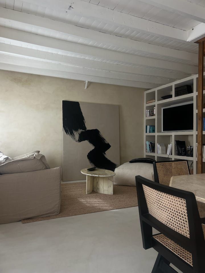 Salon moderne avec un tableau abstrait noir et beige, une étagère pour la télévision, un canapé et un fauteuil en osier sous un plafond mansardé.