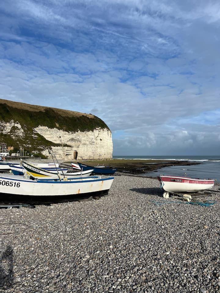 De petites embarcations sur une plage de galets, au pied de falaises de craie blanche, sous un ciel bleu nuageux.