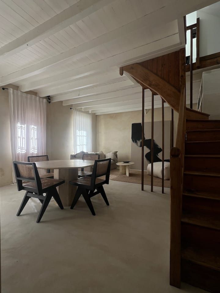 Salon lumineux avec table à manger, chaises, grandes fenêtres et escalier en bois