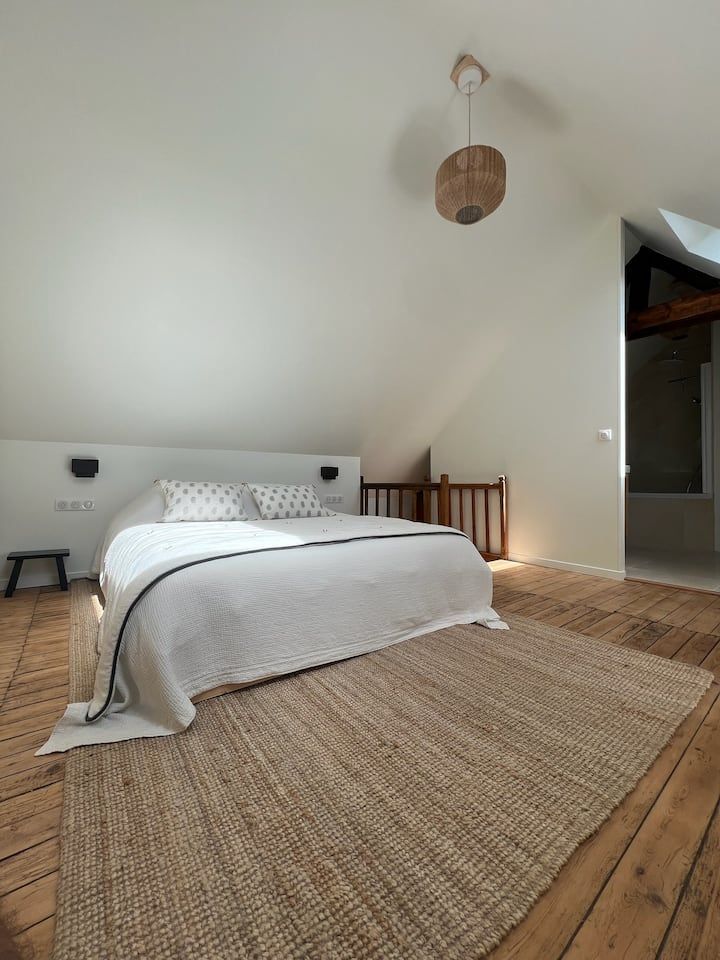 Chambre mansardée lumineuse avec un lit bas, un tapis tissé, des murs blancs et une suspension lumineuse.