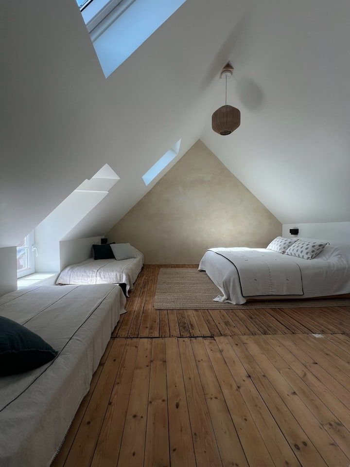 Chambre mansardée minimaliste avec murs blancs, puits de lumière, trois lits et une suspension lumineuse en tissu tressé.