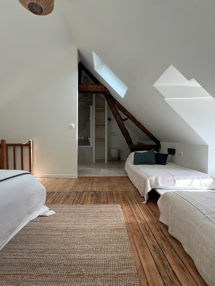 Chambre mansardée lumineuse avec murs blancs en pente, puits de lumière, parquet, tapis tissé et deux lits bas blancs.