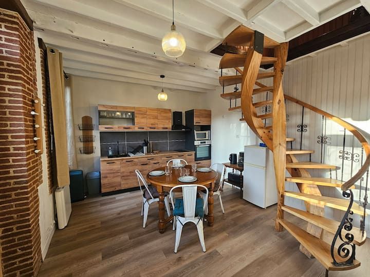 Coin repas cosy en mezzanine avec escalier en colimaçon en bois, mur de briques et coin cuisine sous poutres apparentes.