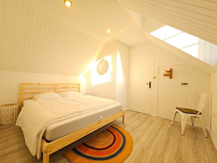Chambre mansardée lumineuse avec un lit en bois, un tapis orange, un puits de lumière et une chaise blanche près de la porte.