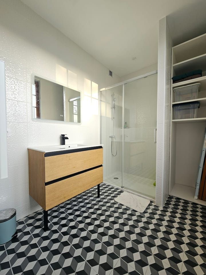 Salle de bains moderne avec sol à motifs noirs et blancs, meuble-lavabo en bois, douche en verre et étagères ouvertes.