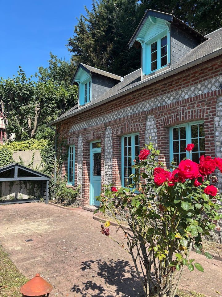 Maison en briques avec des boiseries turquoise et des lucarnes, des roses rouges devant et une cour en terre battue sous un ciel bleu.