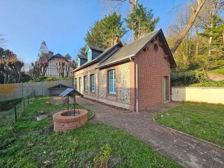 Maisonnette en briques avec fenêtres à encadrement turquoise, un puits en briques devant et un chemin de jardin dans une cour ensoleillée.
