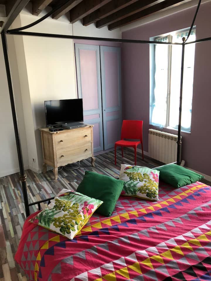 Chambre confortable avec un couvre-lit coloré, une chaise rouge, une télévision sur la commode et une grande fenêtre avec poutres apparentes.