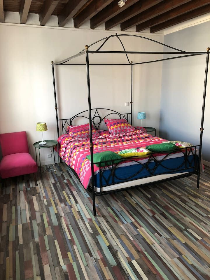 Chambre avec un lit à baldaquin noir, une couette colorée et une moquette à rayures multicolores sous des poutres en bois apparentes.