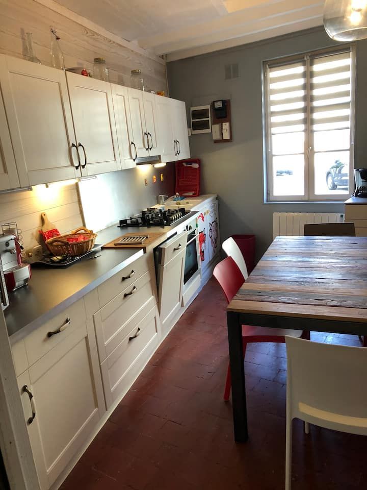 Cuisine lumineuse avec placards blancs, table en bois, chaises rouges et fenêtre avec stores.