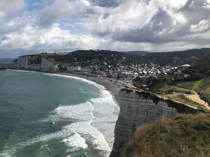 Ville côtière à flanc de falaise surplombant une plage incurvée et une mer turquoise sous un ciel nuageux
