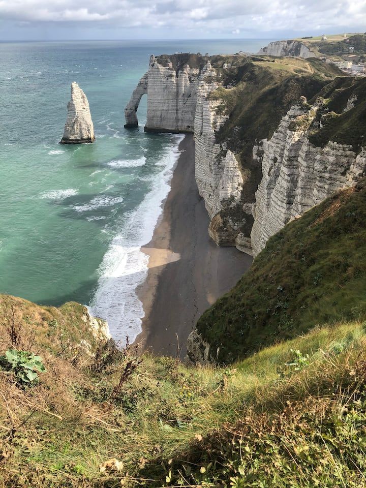 Côte bordée de falaises avec un rocher isolé, une plage de galets noirs et de l'herbe verte sous un ciel nuageux