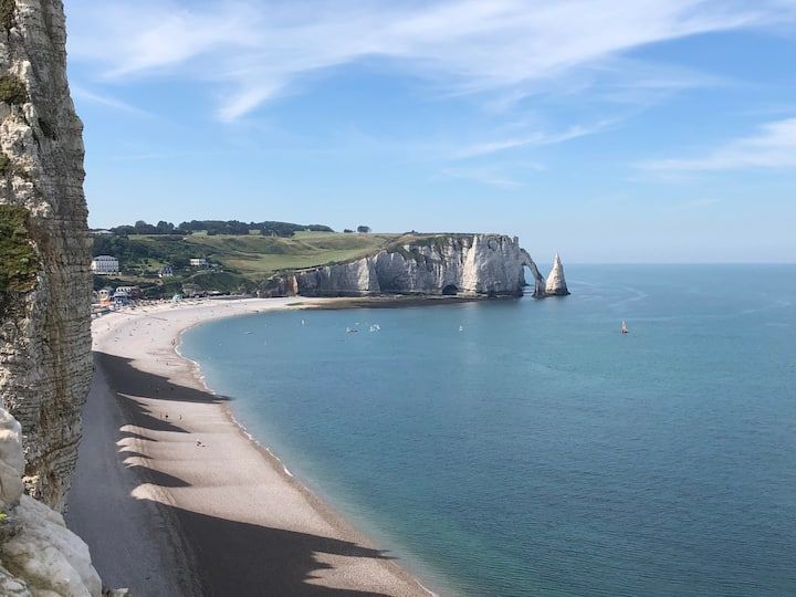 Vue du haut d'une falaise sur une plage de sable fin et une baie bleue calme, bordée de falaises de craie blanche et d'un voilier.