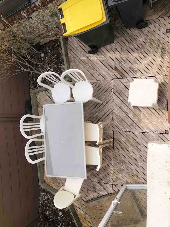Terrasse extérieure en bois avec table et chaises de salle à manger blanches, poubelle à couvercle jaune et tabouret carré.