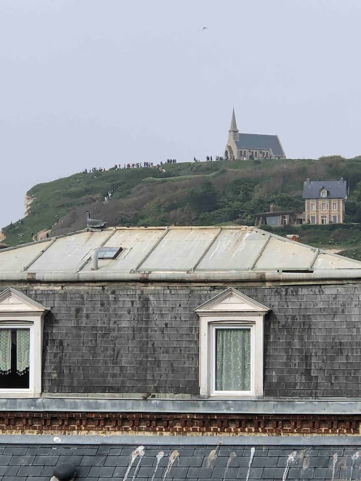 Toits de pierre au premier plan, avec une église sur une colline verdoyante sous un ciel gris.