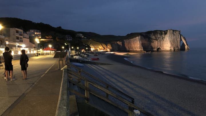 Promenade nocturne en bord de mer, avec des gens marchant le long d'une plage illuminée et des falaises au loin.