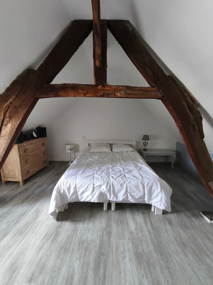 Chambre mansardée avec poutres apparentes, un lit centré en dessous et une commode sur la gauche.