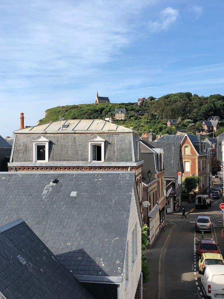 Vue sur des toits d'ardoise donnant sur une rue étroite, avec une colline verdoyante et une petite église sous un ciel bleu.