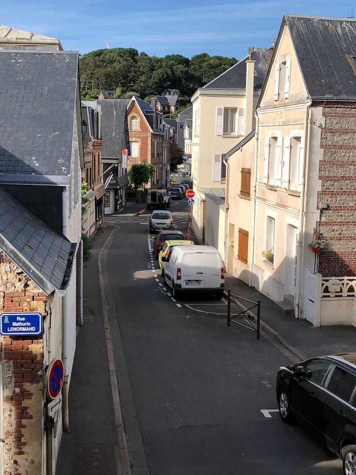 Rue étroite bordée de voitures garées et de bâtiments aux couleurs pastel, dans une ville côtière ensoleillée.