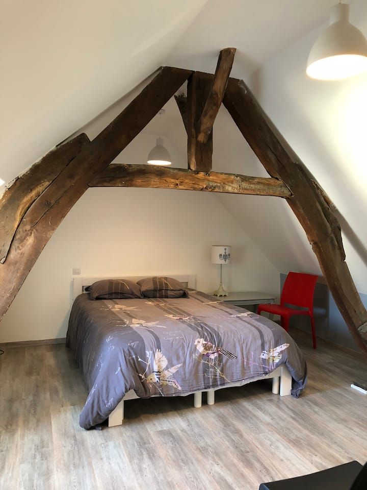 Chambre mansardée rustique avec poutres apparentes, un lit et une chaise rouge sous des murs blancs en pente.