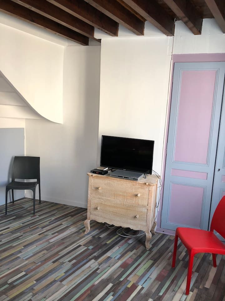 Petite pièce avec une télévision sur une commode en bois, deux chaises et une porte rose, avec des poutres apparentes sombres au plafond.
