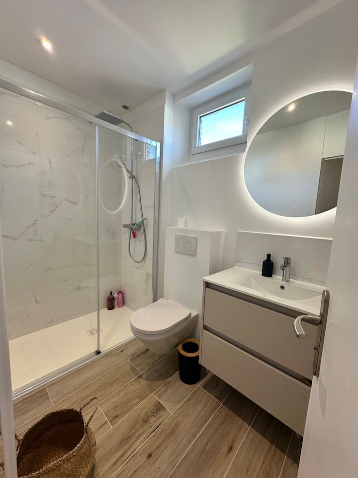 Salle de bains moderne avec douche en verre, toilettes suspendues, meuble-lavabo, miroir rond et sol imitation bois.