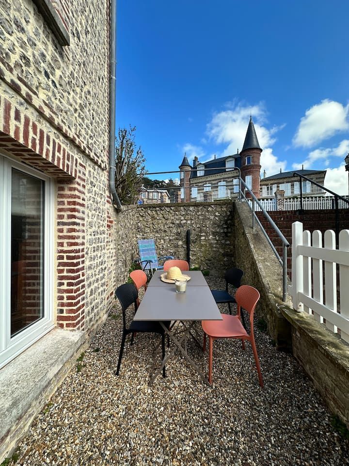 Petite terrasse en gravier avec une table, des chaises colorées et un muret en pierre jouxtant un bâtiment en briques et une clôture blanche.