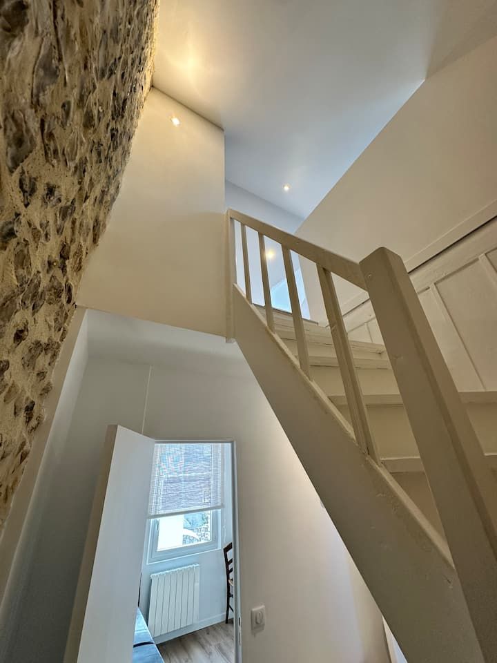 Escalier étroit aux murs beiges, rampe blanche et petite fenêtre au-dessus d'une porte.