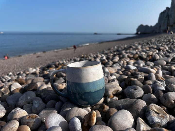 Mug en céramique bleue sur une plage de galets avec l'océan et des falaises en arrière-plan