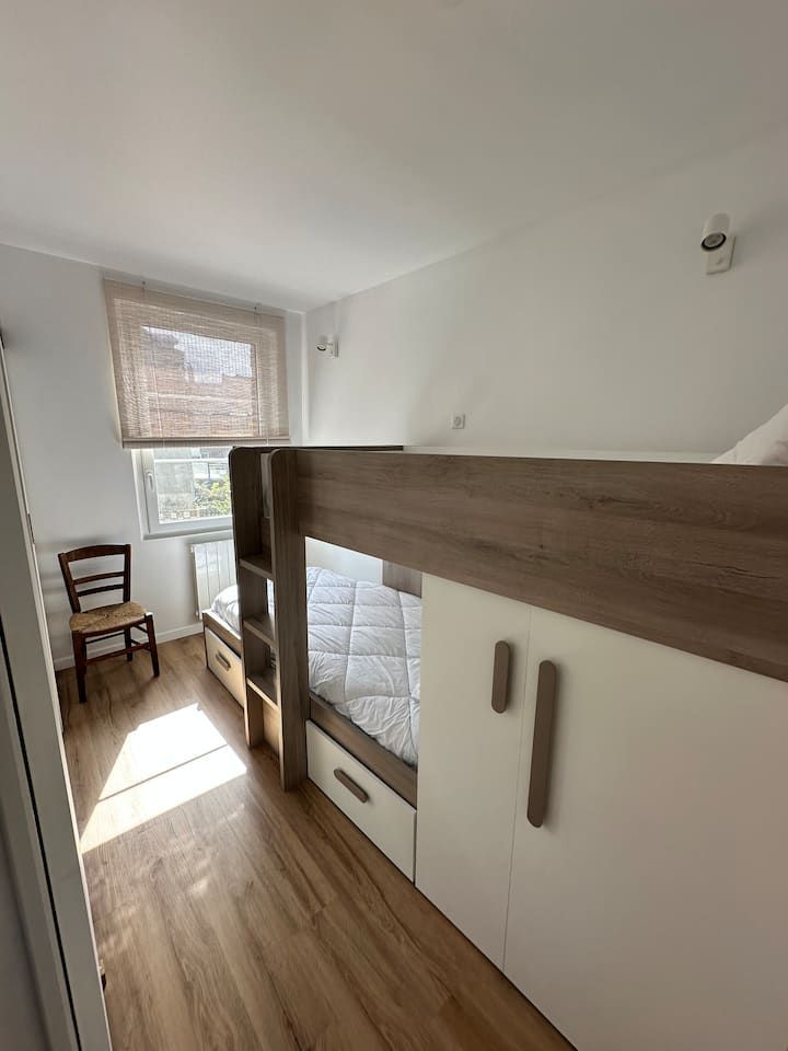 Petite chambre lumineuse avec lit mezzanine, parquet, murs blancs et fenêtre laissant entrer la lumière du soleil.