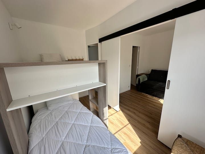 Petite chambre avec un lit mezzanine, des étagères blanches et une porte ouverte donnant sur un couloir desservant une autre pièce.